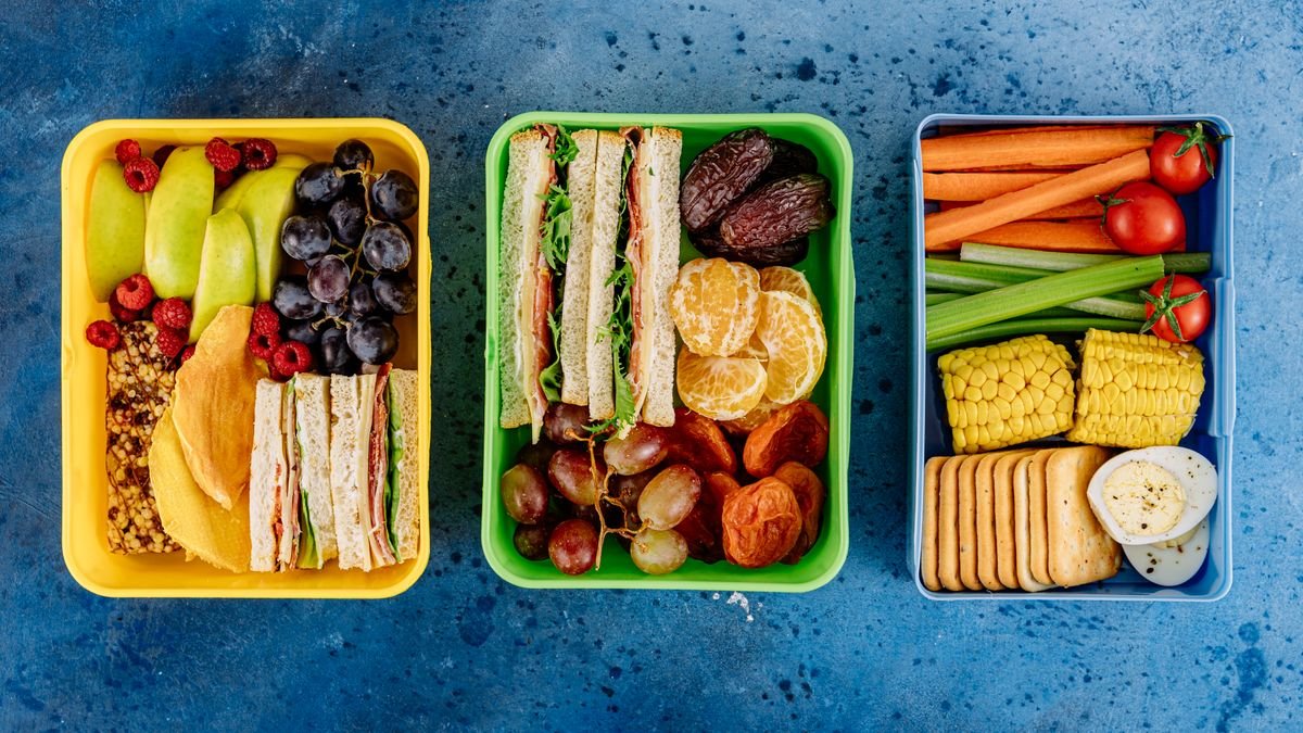 Três lancheiras escolares abertas com frutas, sanduíches e legumes sobre mesa azul