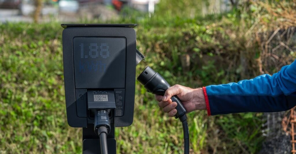 Mão segura o conector de um carregador de carro elétrico ao lado do totem de recarga