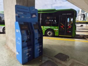 Dois totens azuis de autoatendimento do Cartão Mobilidade em terminal do DF, com ônibus verde ao fundo