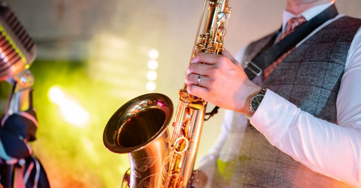 Closeup de saxofone dourado tocado por músico de colete, ao lado de microfone vintage