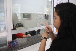 Mulher se comunica em Libras diante de um guichê de atendimento enquanto um celular apoiado no balcão exibe a videochamada com o intérprete