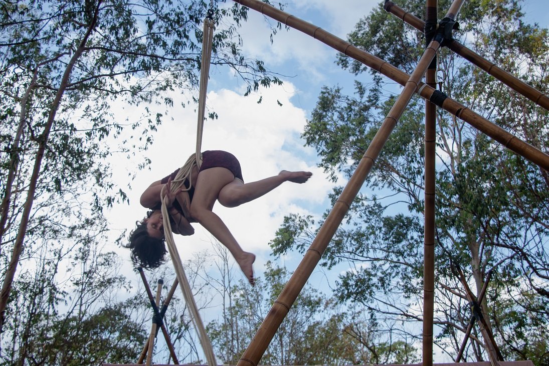 Acrobata suspensa em cordas presas a uma estrutura de bambu, com o céu e as árvores do Cerrado ao fundo