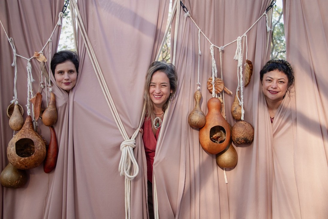 Três artistas do espetáculo entre cortinas cor de areia, cercadas por cabaças penduradas em cordas