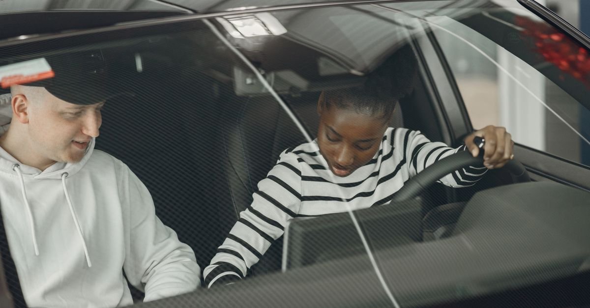 Mulher ao volante de um carro com as duas mãos no aro, ao lado de um instrutor no banco do carona durante aula de direção
