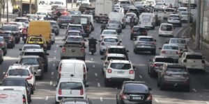 Trânsito congestionado em avenida brasileira com carros, vans e motociclistas entre as faixas