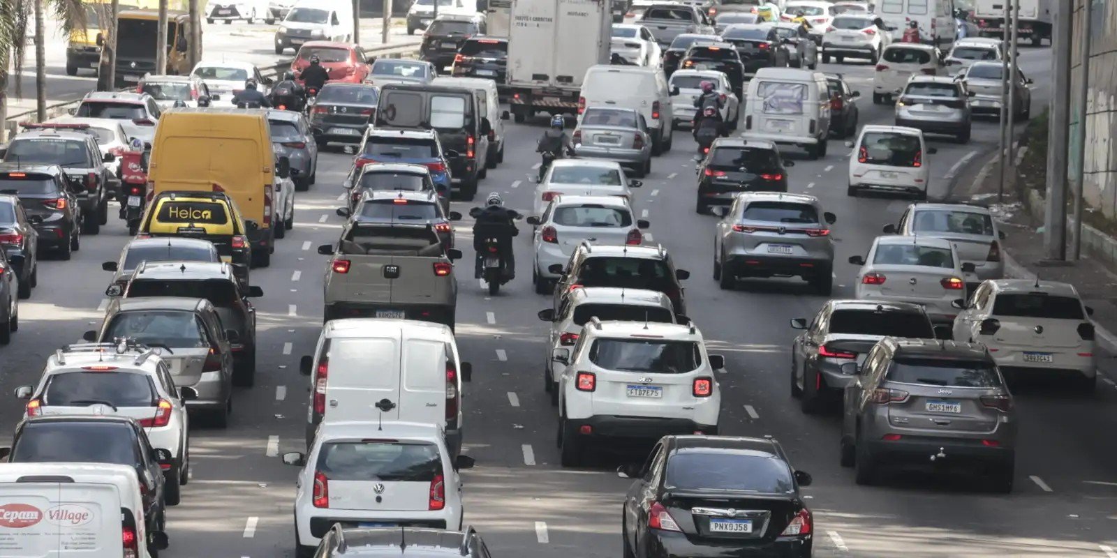 Trânsito congestionado em avenida brasileira com carros, vans e motociclistas entre as faixas