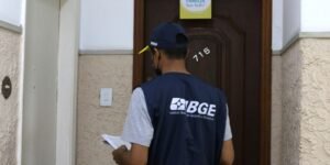 Recenseador com colete do IBGE em frente à porta de um apartamento durante a coleta de dados do Censo