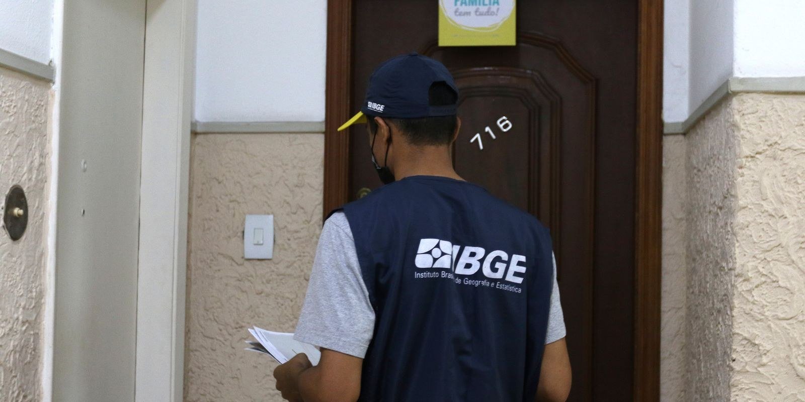 Recenseador com colete do IBGE em frente à porta de um apartamento durante a coleta de dados do Censo