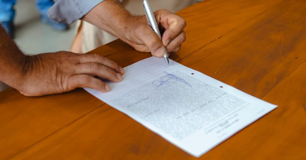 Mãos de uma pessoa assinando um documento com caneta sobre uma mesa de madeira