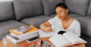 Mulher estuda com cadernos, livros e canetas sobre a mesa, com o celular ao lado