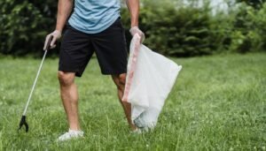 Voluntário de camiseta azul recolhe lixo em gramado com garra coletora e saco plástico branco