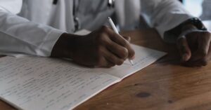 Médico de jaleco branco e estetoscópio escreve à mão em um caderno apoiado sobre mesa de madeira