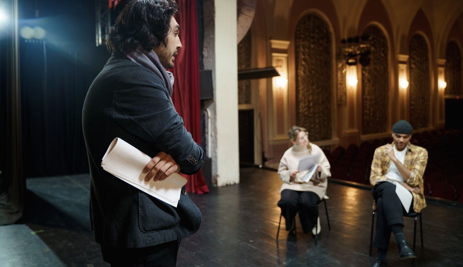 Ator de casaco escuro segura texto impresso em palco de teatro enquanto dois colegas sentados acompanham o ensaio