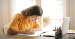 Estudante escreve em caderno ao lado de notebook sobre mesa de madeira
