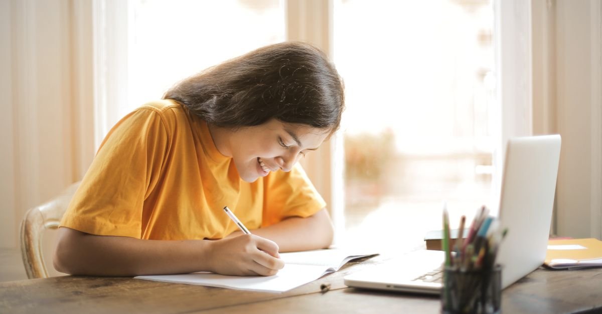 Estudante escreve em caderno ao lado de notebook sobre mesa de madeira