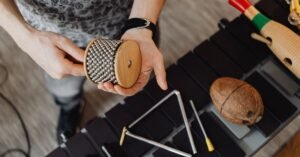 Mãos segurando instrumento de percussão de madeira sobre mesa com triângulo e coco