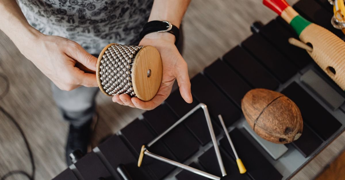 Mãos segurando instrumento de percussão de madeira sobre mesa com triângulo e coco