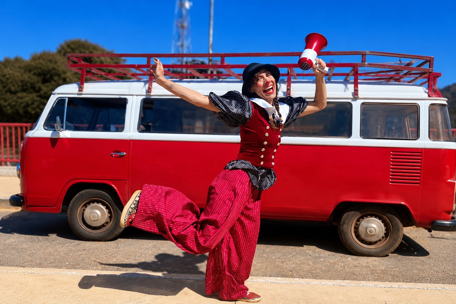 Palhaça de colete vermelho e calça listrada ergue um megafone diante de uma kombi vermelha, ao sol