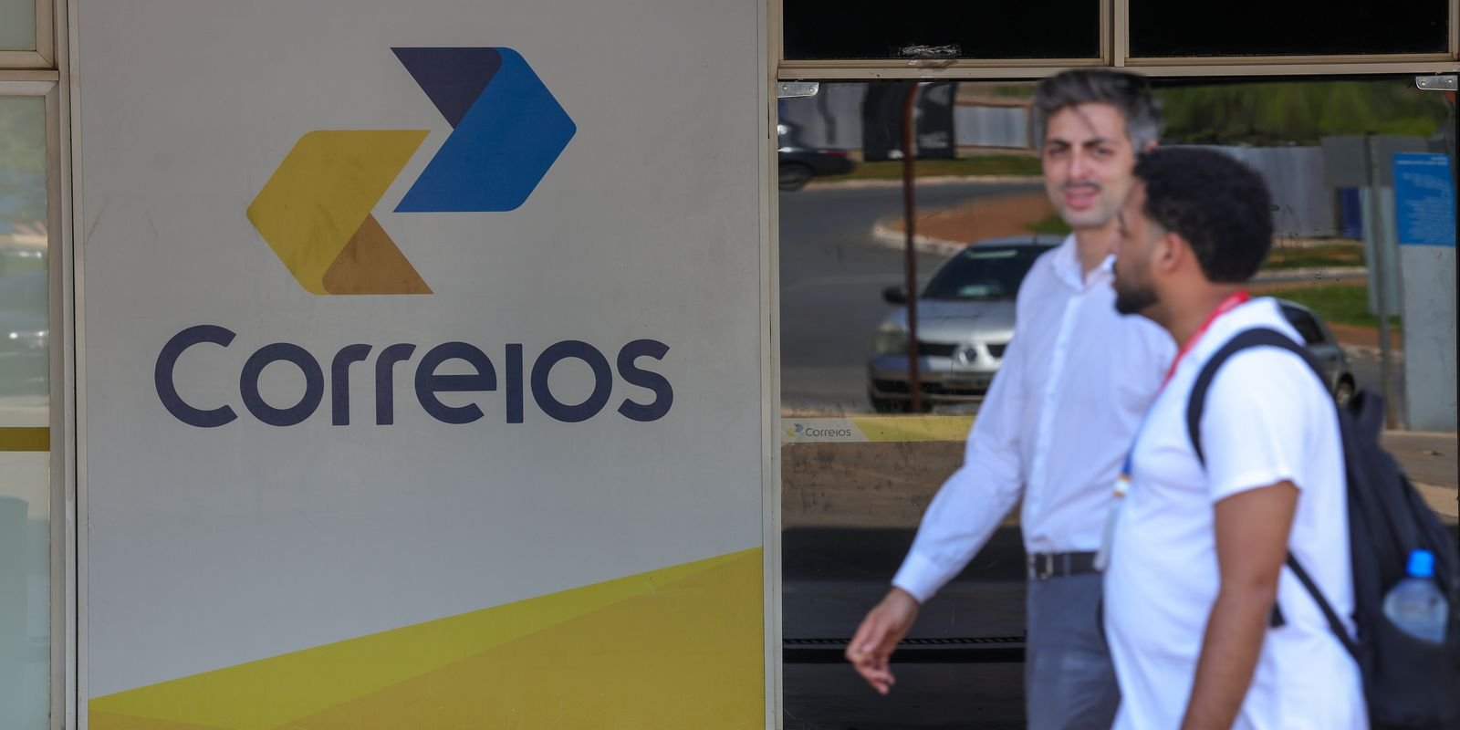 Fachada de agência dos Correios com o logotipo da empresa e dois homens passando na calçada