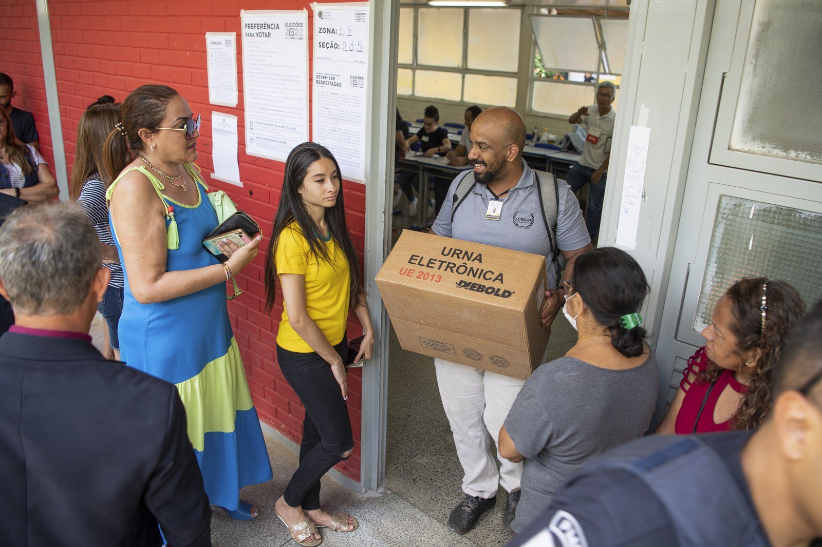 Fila de eleitores na entrada de uma seção eleitoral ao lado de servidor que carrega caixa de urna eletrônica