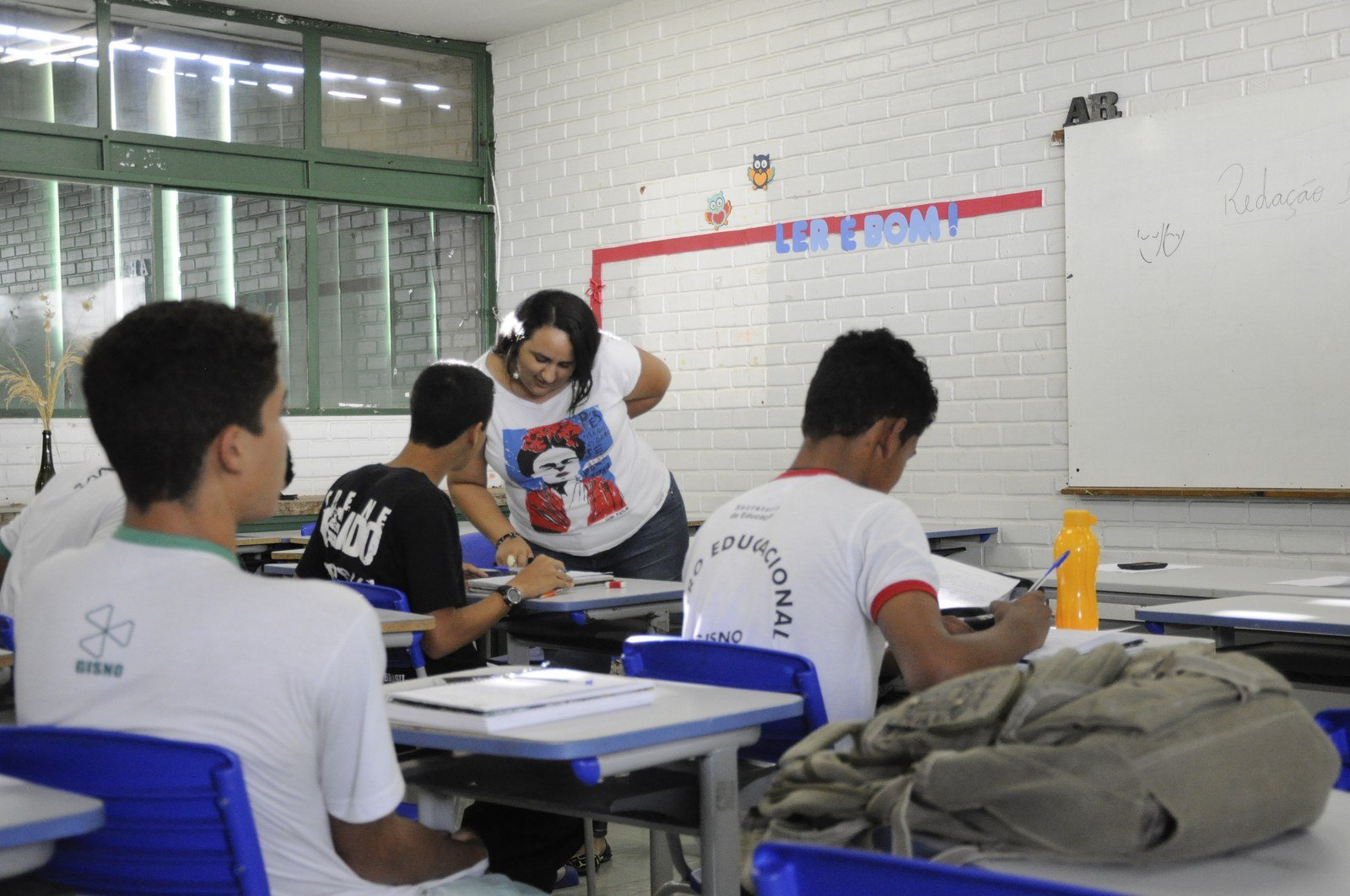 Professora atende aluno em sala de aula de escola pública do Distrito Federal, com colegas de uniforme escrevendo nas carteiras