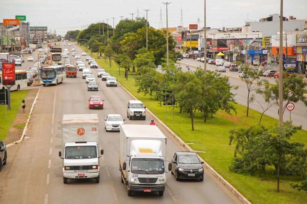 Avenida movimentada em Taguatinga, no Distrito Federal, com ônibus, caminhões e comércio nas laterais
