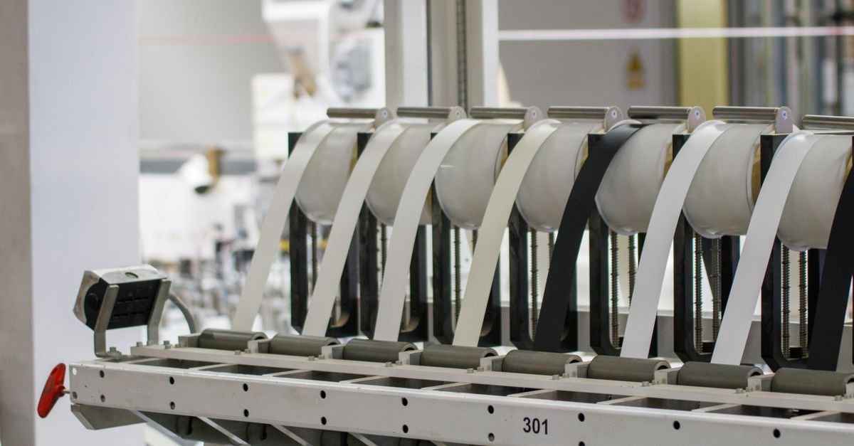 Máquina industrial em operação numa linha de produção, com bobinas de fita branca e preta