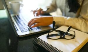 Mãos digitam em notebook ao lado de caderno e óculos sobre a mesa