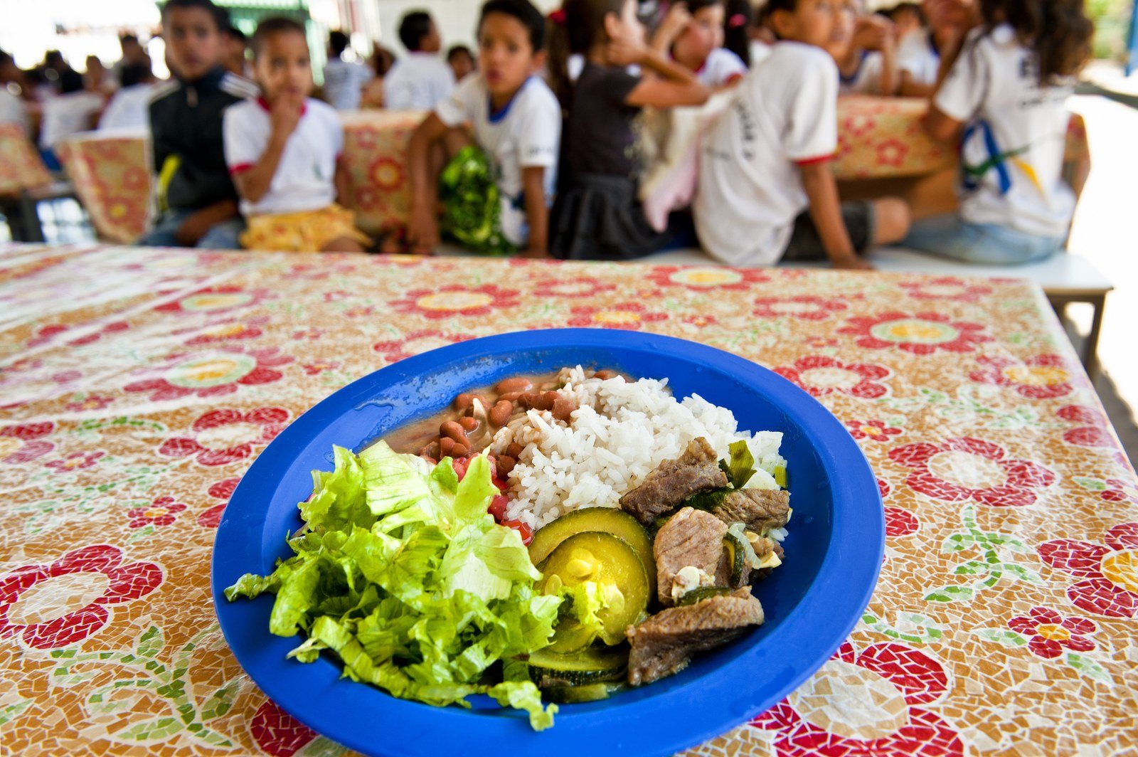 Prato azul com arroz, feijão, carne, abobrinha e salada sobre mesa de escola pública, com estudantes ao fundo