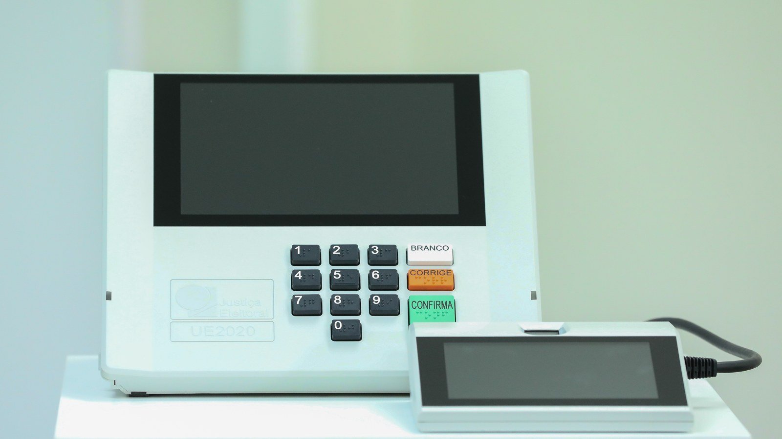 Urna eletrônica modelo UE2020 da Justiça Eleitoral, com teclado numérico e botões BRANCO, CORRIGE e CONFIRMA, ao lado do terminal do mesário