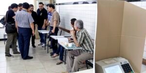 Mesários sentados à mesa receptora de votos ao lado de cabina com urna eletrônica da Justiça Eleitoral