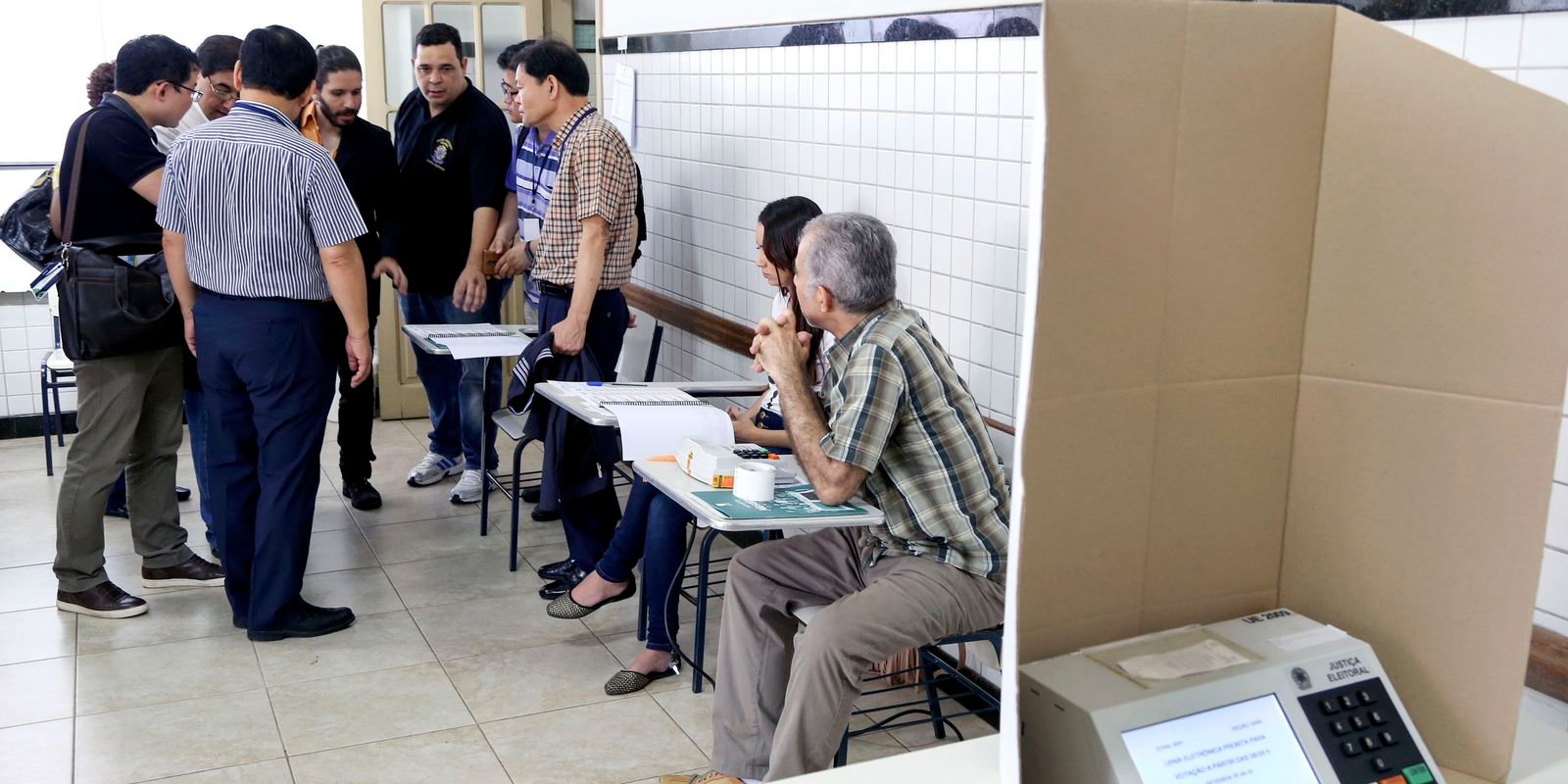 Mesários sentados à mesa receptora de votos ao lado de cabina com urna eletrônica da Justiça Eleitoral