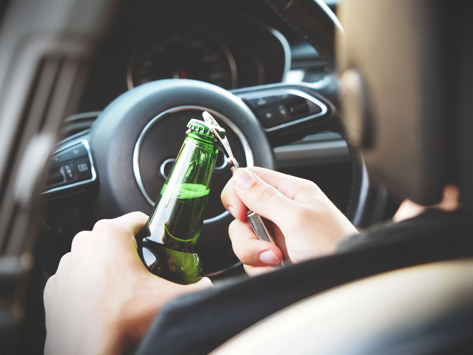 Mão abre uma garrafa de cerveja ao volante de um carro, com o painel e o volante ao fundo