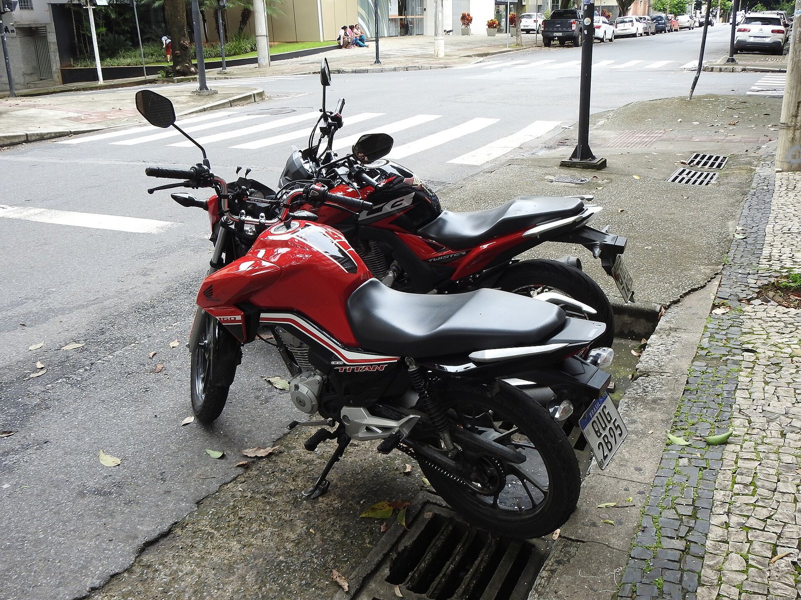 Duas motocicletas Honda vermelhas estacionadas na guia de uma rua, a da frente uma CG 160 Titan com placa brasileira