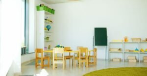 Sala de educação infantil vazia com mesa e cadeiras pequenas, prateleiras com materiais pedagógicos