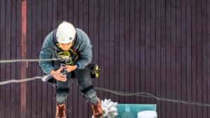 Trabalhador com capacete e cinturão de segurança tipo paraquedista suspenso por cordas em fachada