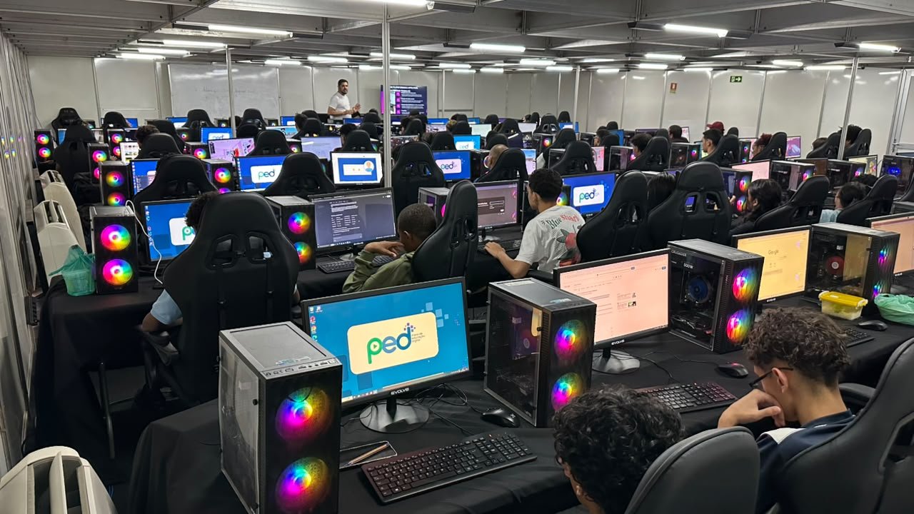 Sala de aula do PED com dezenas de alunos diante de computadores, com a marca do projeto nas telas