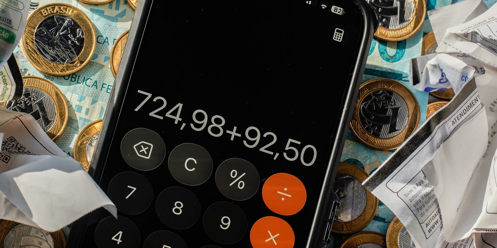 Celular com calculadora aberta sobre moedas de real, cédulas e notas fiscais amassadas