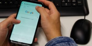 Mao segura celular com a pagina de busca aberta, ao lado de teclado e mouse sobre a mesa