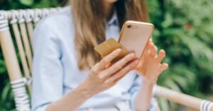 Mulher sentada ao ar livre segura celular e cartão enquanto faz um pagamento pelo aplicativo do banco