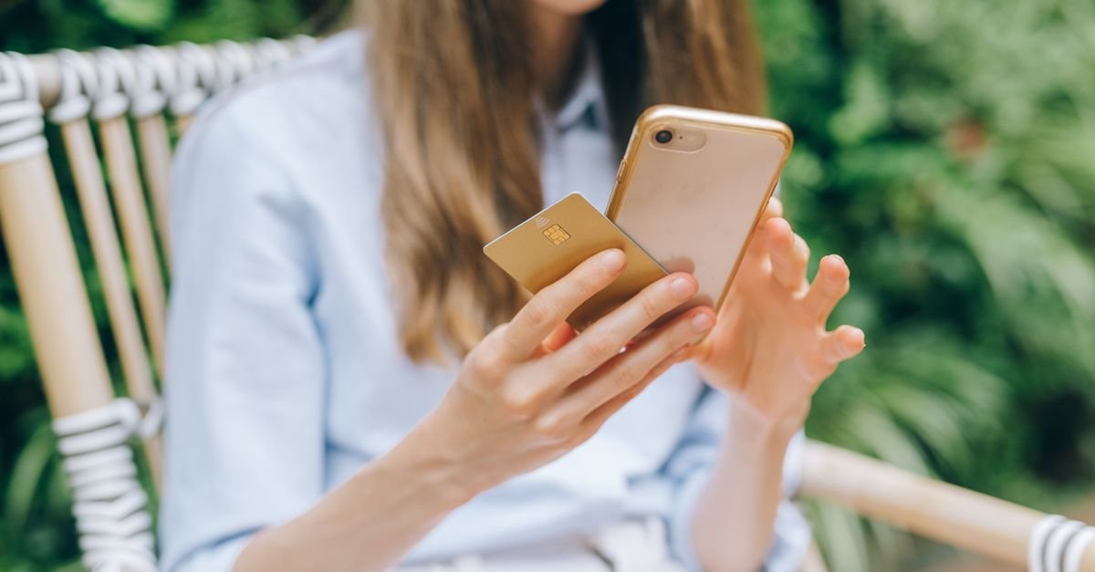 Mulher sentada ao ar livre segura celular e cartão enquanto faz um pagamento pelo aplicativo do banco