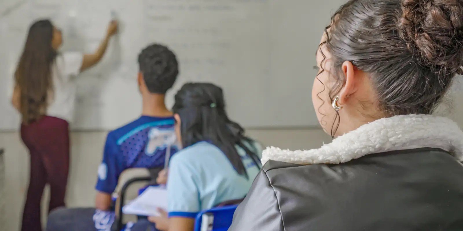 Professora escreve no quadro branco enquanto estudantes acompanham a aula em sala de aula