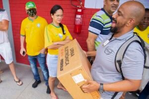 Servidor da Justiça Eleitoral carrega caixa de urna eletrônica diante de fila de eleitores