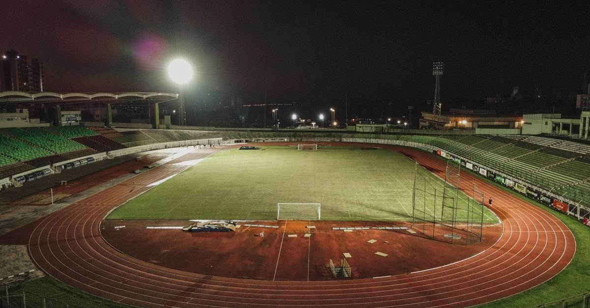 Estádio de futebol iluminado à noite com placas de publicidade ao redor da pista