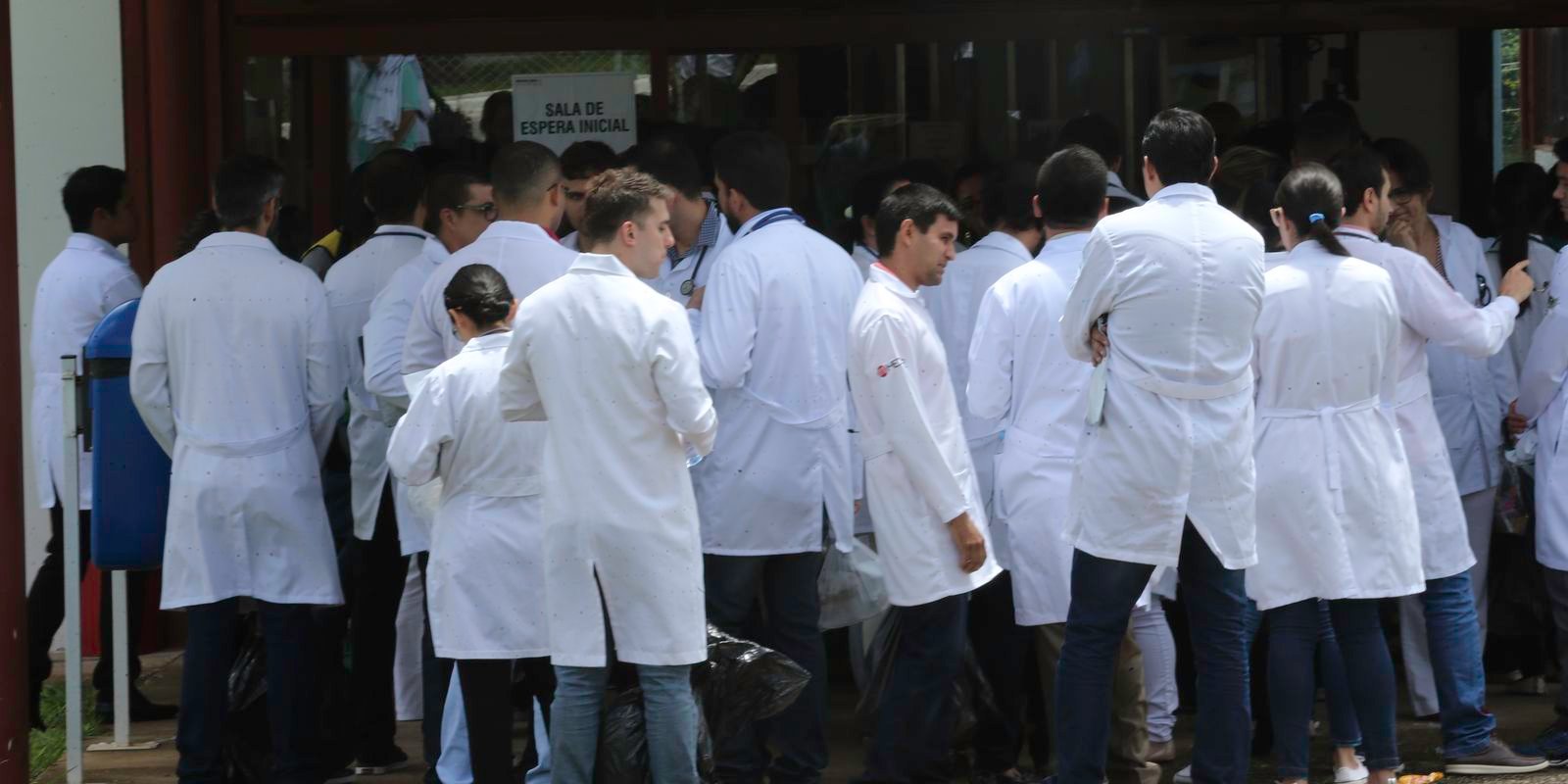Candidatos de jaleco branco em fila na entrada do local de prova de exame para médicos