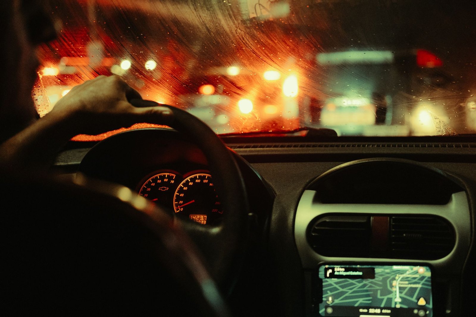 Interior de um carro à noite, com a mão do motorista no volante e o painel aceso