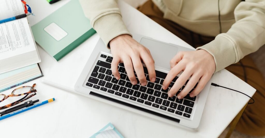 Mãos digitando em notebook sobre mesa com livros e caderno, em referência a curso on-line