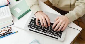Mãos digitando em notebook sobre mesa com livros e caderno, em referência a curso on-line