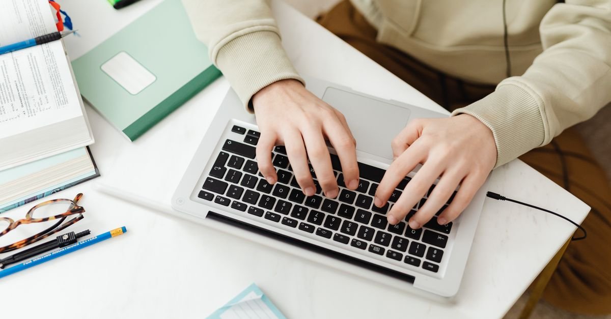 Mãos digitando em notebook sobre mesa com livros e caderno, em referência a curso on-line