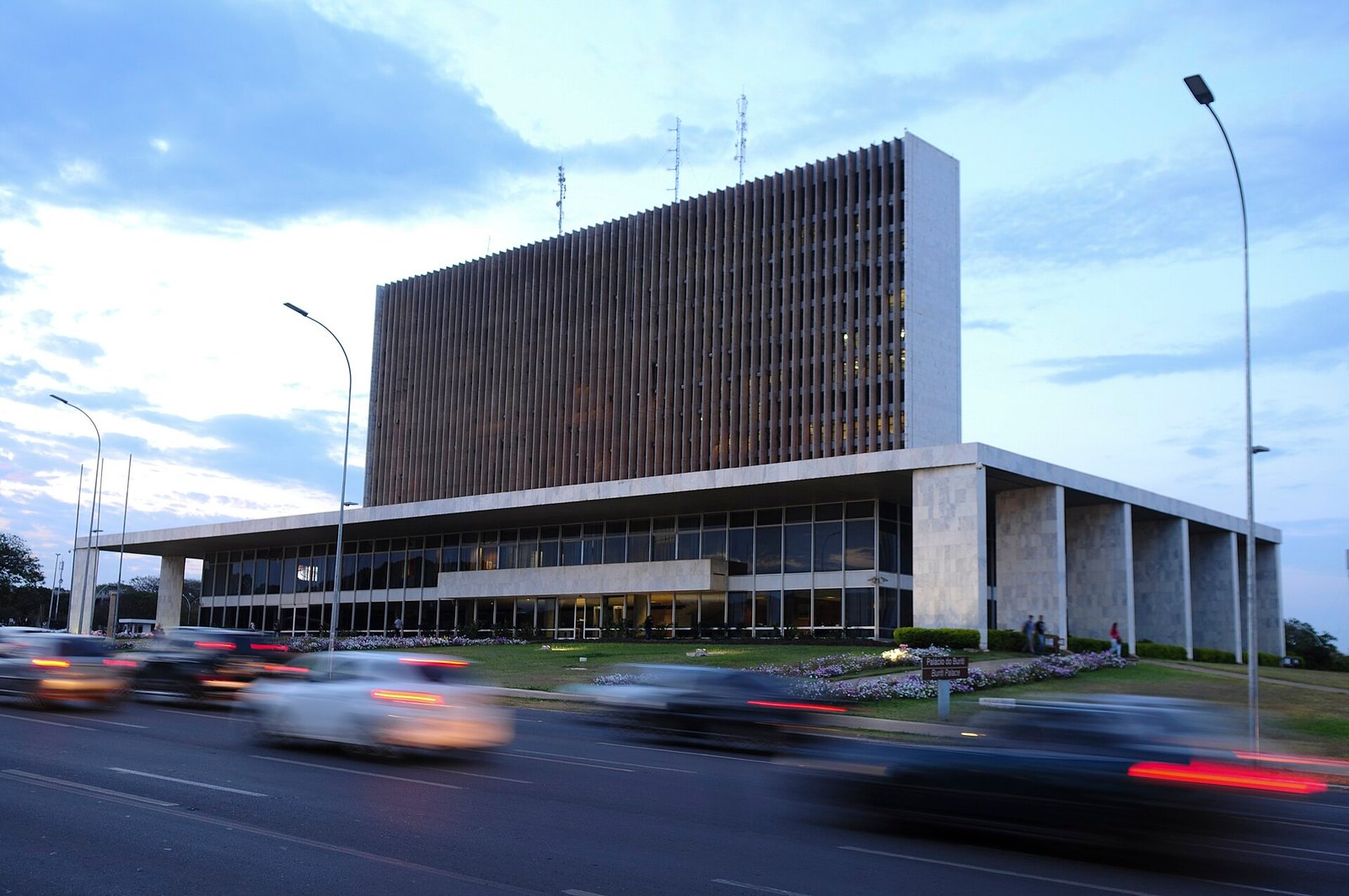 Fachada do Palácio do Buriti, sede do governo do Distrito Federal, ao entardecer, com carros passando na via em primeiro plano
